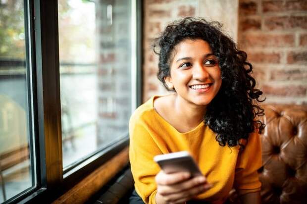 A smiling person sitting on a couch near a window and holding a cell phone.