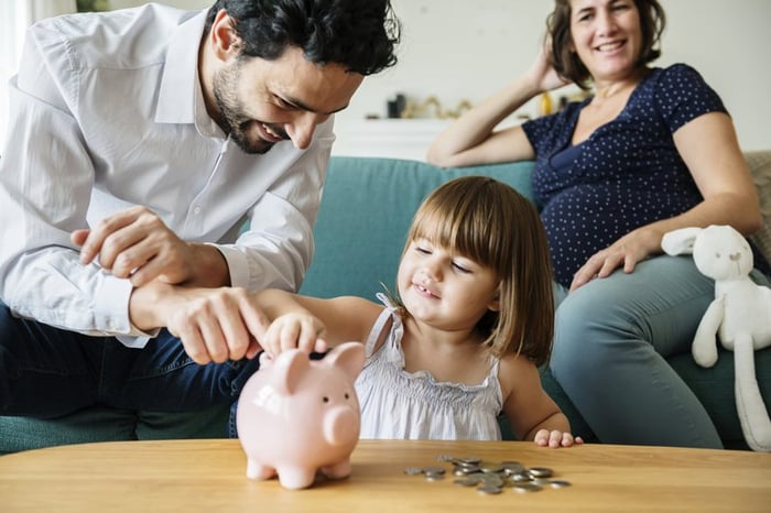 Happy Father Showing His Daughter How To Put Coins In Piggy Bank While Mom Watches
