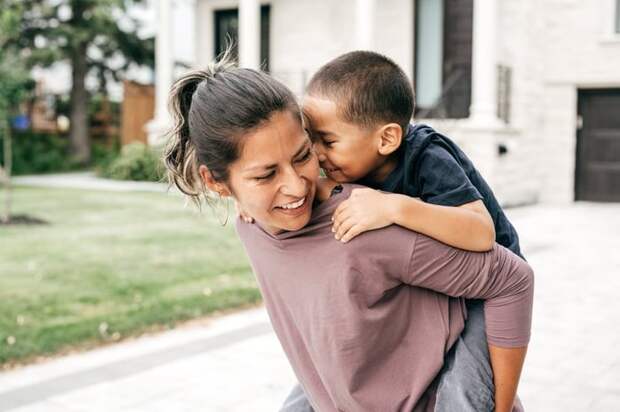 A mom giving her young son a piggyback ride in front of their house.