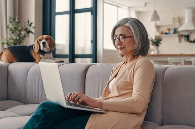 A person using a laptop on a couch with a dog nearby.