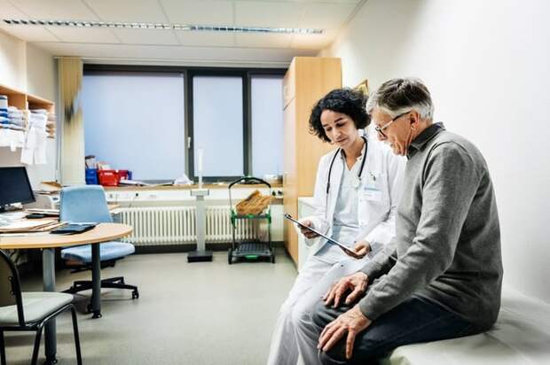 A doctor sitting next to a patient in an exam room and going over test results on a clipboard.