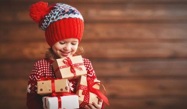 A grinning little girl in a knit cap holding an armful of gifts.