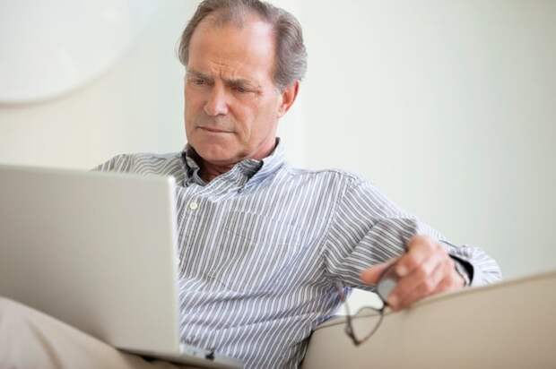 A person seated on a couch holding their glasses while critically reading material on an open laptop.