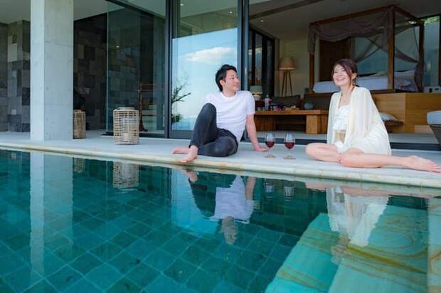 A man and woman sitting at the edge of a pool outside their room in a luxury hotel.