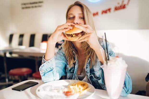 A person eating a hamburger with a plate of fries and a milkshake on the table in a fast food restaurant.