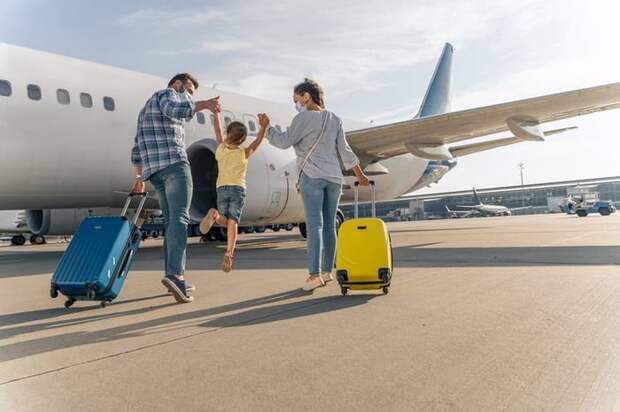 Two parents holding their jumping child's hands between them while rolling their suitcases toward an airplane on the tarmac.