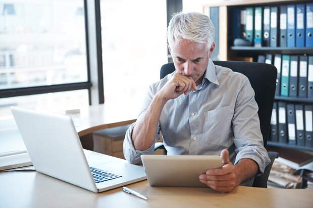 A senior seated at a desk frowns at a tablet.
