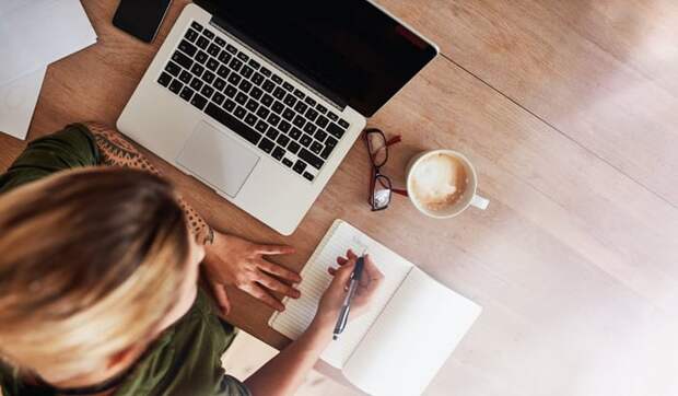 A woman writing out a to-do list at a desk next to a laptop and cup of coffee.