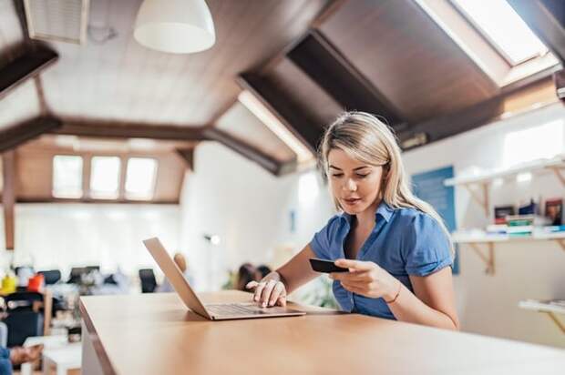 A woman sitting at a table in her home and typing on her laptop while holding her credit card.