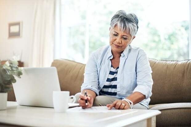 A person sitting on a couch and writing on a piece of paper next to a laptop open on a table.