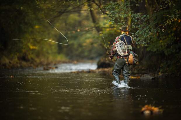 A man stands in a river surrounded by greenery as he makes a cast on the fly