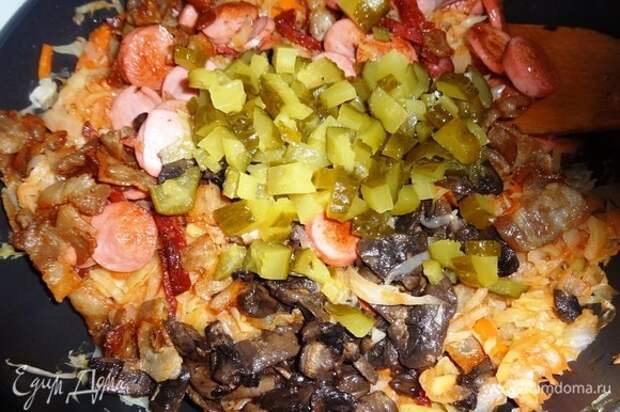 В сковороду с капустой добавить соленые огурцы, обжаренные сосиски, колбасу, бекон, грибы вместе с жиром, перемешать.
