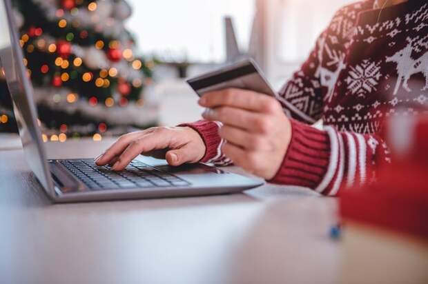 A person in a holiday sweater holding a credit card while typing on a laptop.