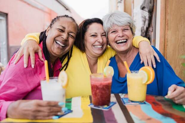 Three friends are seen smiling together, with fruity drinks in front of them.