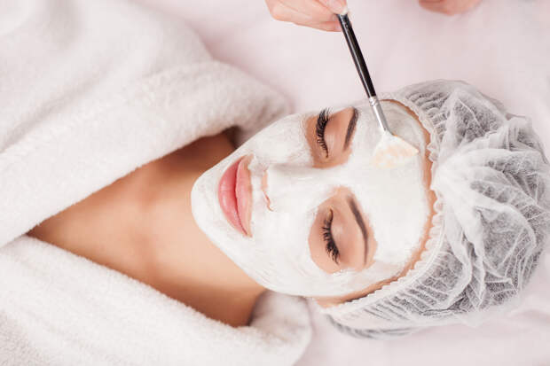 Beautiful young woman is getting facial mask at spa. She is lying and relaxing. Her eyes are closed with pleasure. The cosmetologist is applying cream on her face with brush