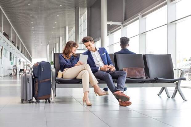 A man and woman sitting at an airport and waiting for their flight while looking at a tablet.