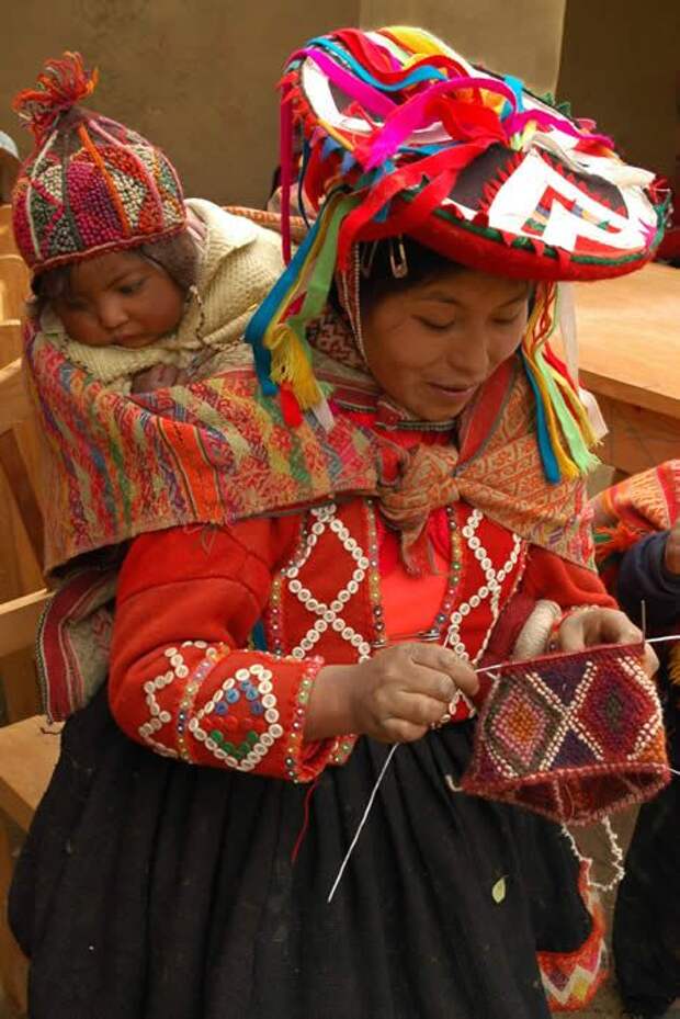 Mother Andean knitter and knitting with baby on back, wearing chullo hand-knit cap ~ Peru