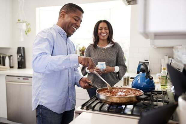 A husband cooking at the stove while his wife looks on, both of them smiling.