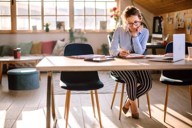 A woman sitting at a desk in a sunny loft and sketching in a notebook next to an open laptop.