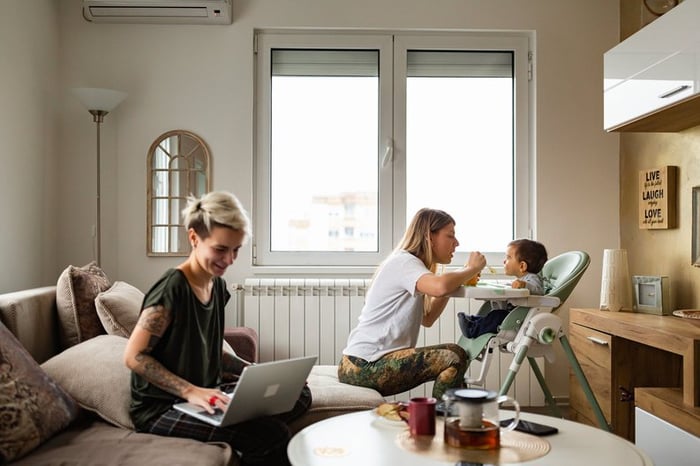 One parent feeding a baby in a high chair while the other works on a laptop on the couch.