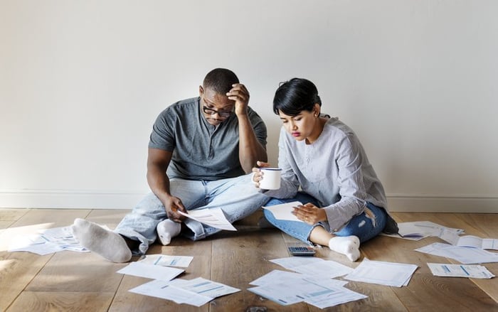A couple sit on the floor in a room with no furniture surrounded by paperwork and bills.