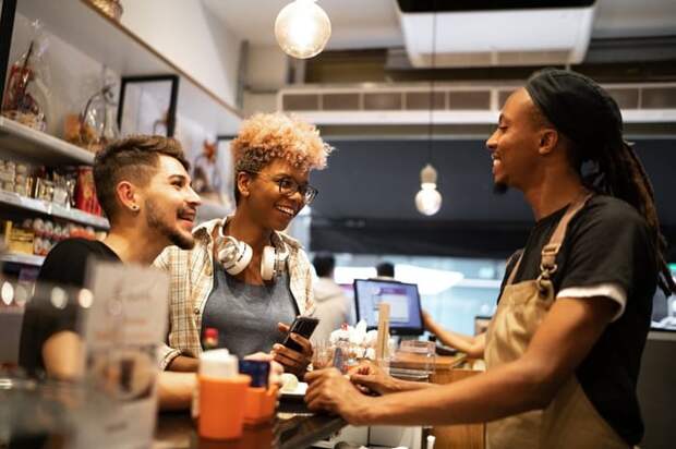 A cafe employee standing behind the counter and happily talking with two smiling customers.