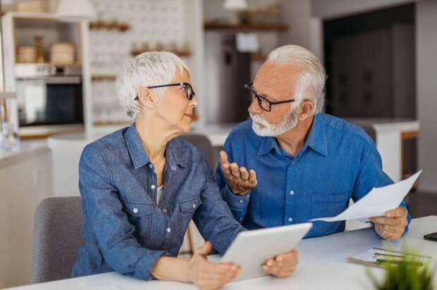 A retired couple sit at a table, engaged in conversation, while the man holds a paper document and the woman holds a digital tablet.