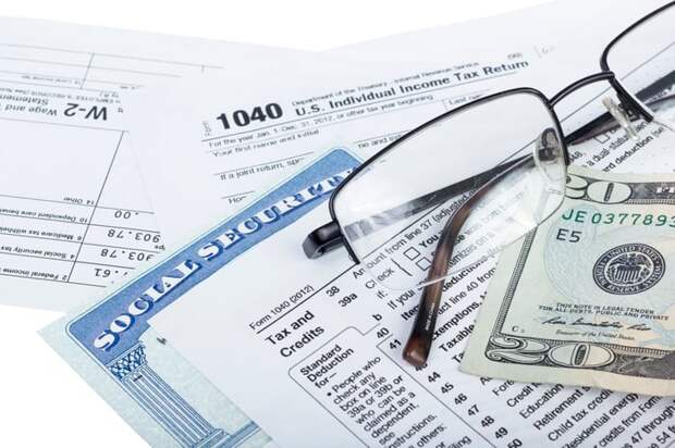 A pair of glasses, a twenty dollar bill, and a Social Security card, set atop federal income tax forms. 