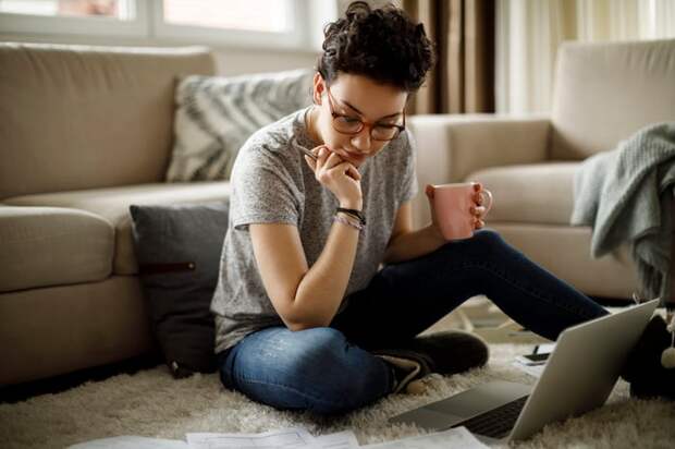 A person holding a mug while sitting on a living room floor and looking at paperwork.