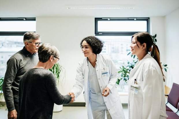 Two older people shaking hands with two smiling doctors in a sunny hospital room.