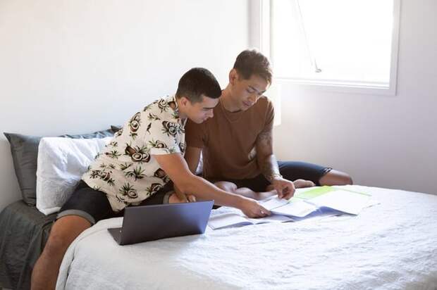 Two people sitting on a bed and looking at paperwork next to an open laptop.