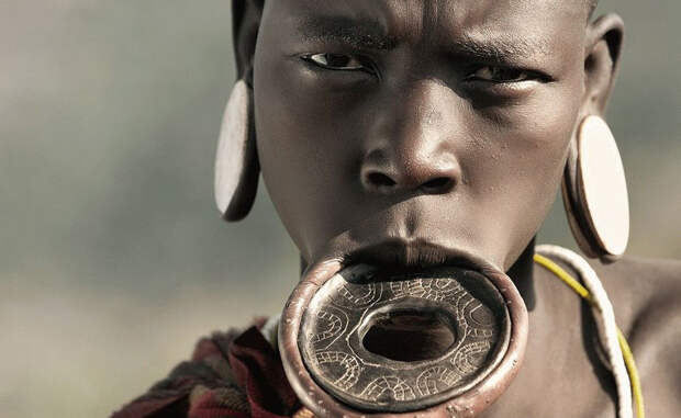 Мурси Африка Для девушек из африканского племени Мурси детство заканчивается примерно в 16 лет. После обряда инициации, шаман разрезает нижнюю губу несчастной и вставляет в отверстие небольшую палочку. Спустя какое-то время девушка начинает носить в губе небольшой каменный диск, постепенно расширяя отверстие. Особо влюбленные в современные тренды дамы мурси умудряются носить в губе настоящие блюда, пятнадцати сантиметров в диаметре. Нижние зубы при этом необходимо удалить, с ними украшение просто не помещается во рту.
