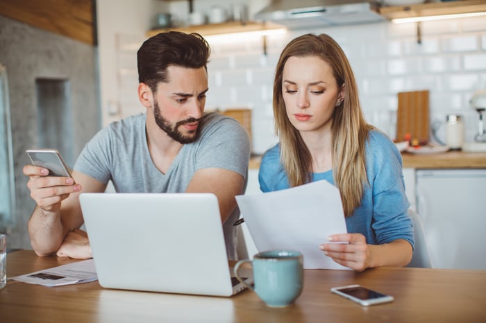 Two people in kitchen looking at laptop and papers.