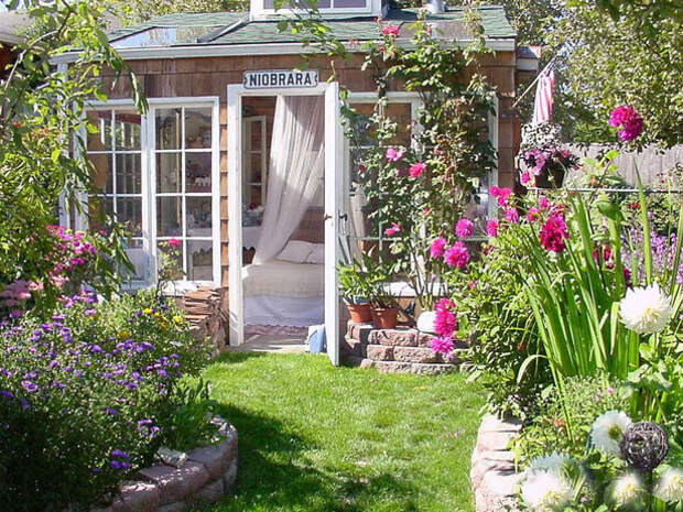 Garden house with canopy style bed inside, colorful flowers in the manicured landscape, and long, tall windows around the front of the garden house.